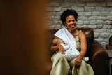 Woman holding a baby wrapped in a white blanket, sitting on a brown leather couch with a brick wall background.